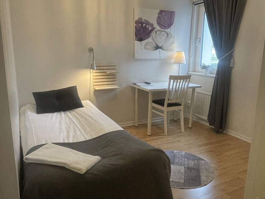 A bedroom with a bed, a desk, and a chair. There is a chandelier above the desk and a window with curtains.