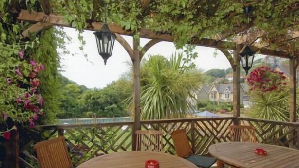 Una terrazza esterna con tavoli e sedie in legno, circondata dal verde e affacciata su case sullo sfondo.