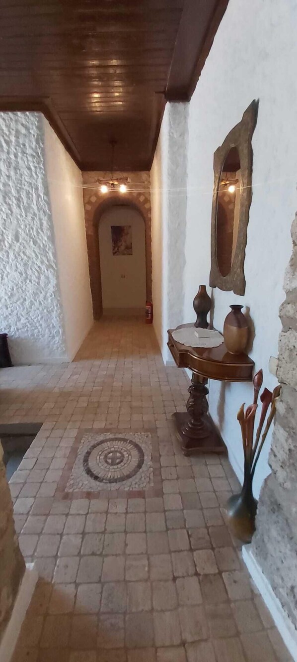 Um corredor estreito com piso estampado, um banco de madeira, um espelho fixado na parede e uma pequena mesa com objetos decorativos.