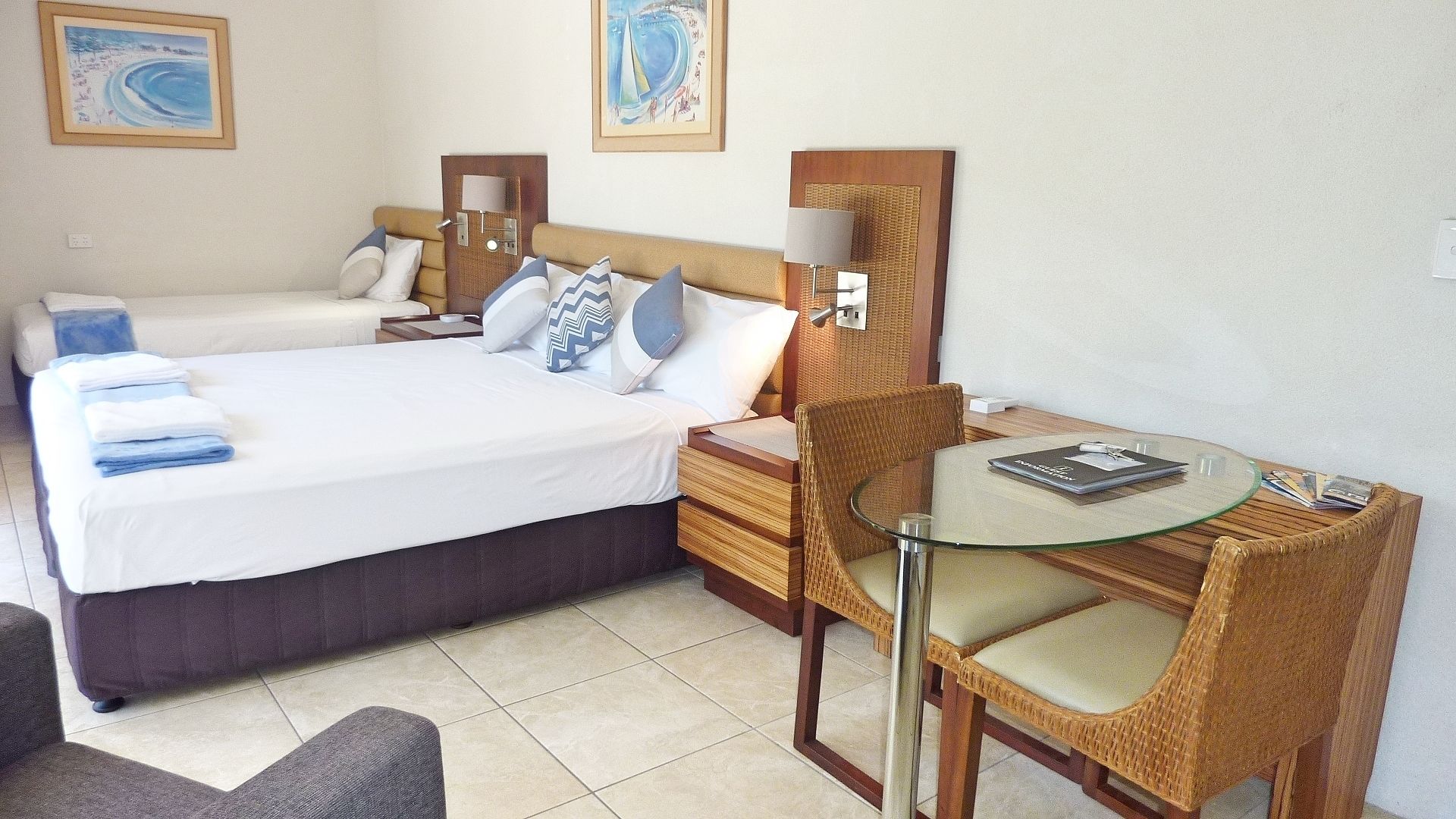 A hotel room with two beds, a glass table, wicker chairs, and artwork on the walls.