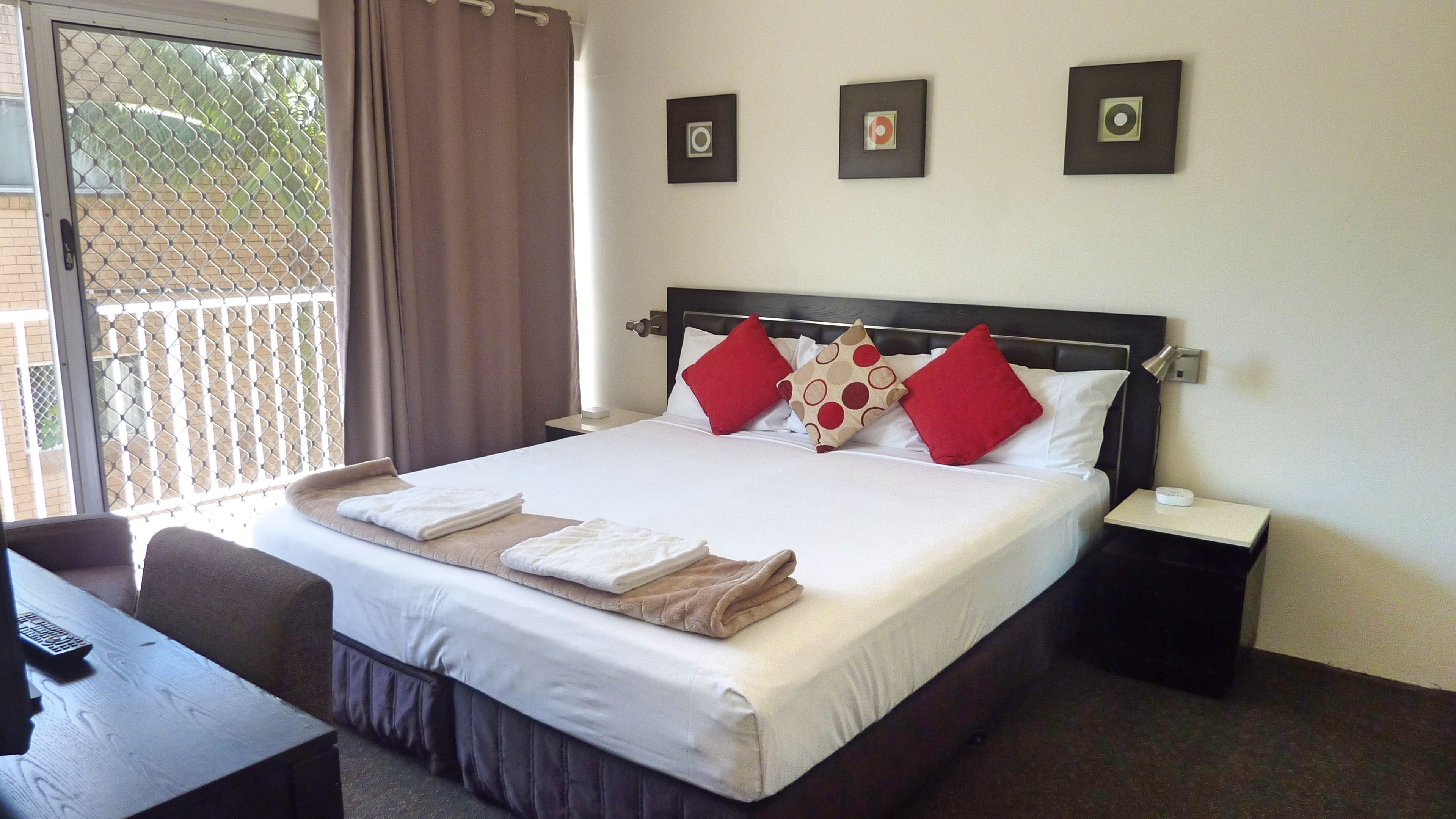 Una cama bien hecha con sábanas blancas y almohadas rojas y beige, una mesita de noche con una lámpara, y vistas a un balcón a través de una ventana con cortinas.