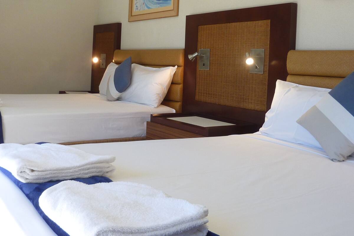 Habitación de hotel con dos camas, cada una con ropa blanca y almohadas azules. Hay toallas sobre las camas y un cabecero de madera con un panel tejido.