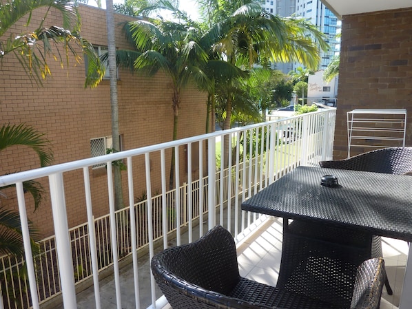 A balcony with wicker furniture, a white railing, and a view of a brick building and palm trees.