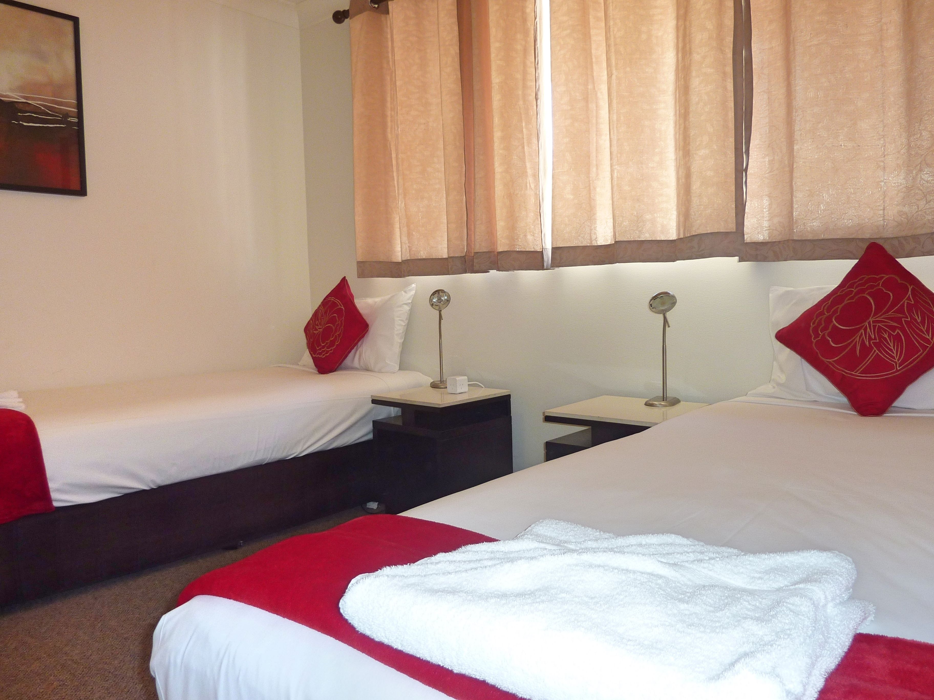 A hotel room with two single beds, each with red and white bedding, a small bedside table with a lamp, and a large bed with white bedding and a red pillow.