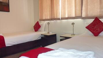 A hotel room with two single beds, each with red and white bedding, a small bedside table with a lamp, and a large bed with white bedding and a red pillow.