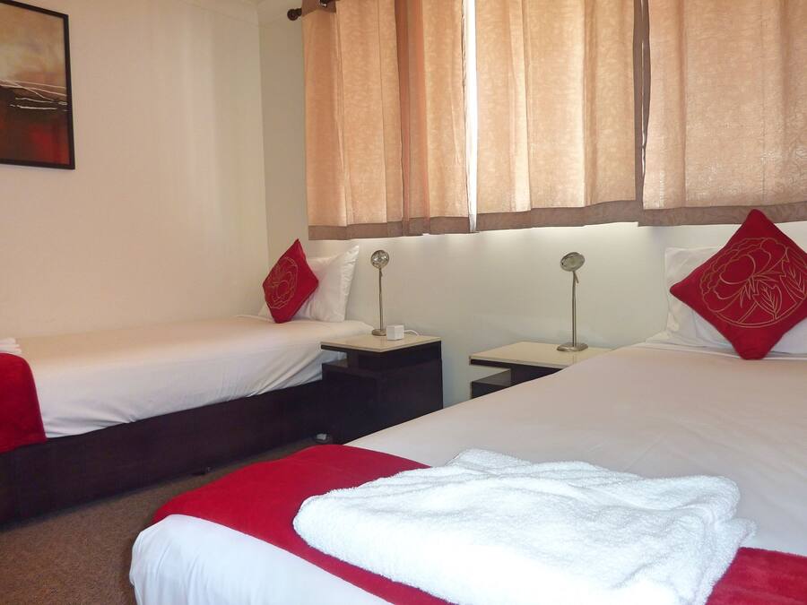 A hotel room with two single beds, each with red and white bedding, a small bedside table with a lamp, and a large bed with white bedding and a red pillow.