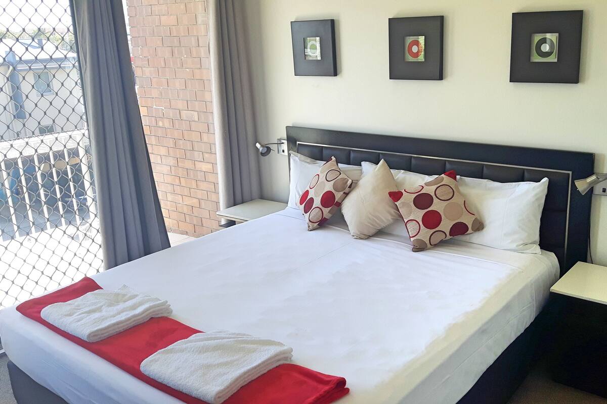 Una cama bien tendida con ropa de cama blanca y roja, dos almohadas con estampado de lunares, una mesita de noche pequeña y una mesita auxiliar.