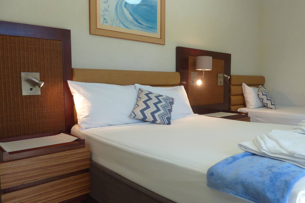 Habitación de hotel con dos camas, cada una con ropa de cama blanca y toallas azules. Presenta cabeceros de madera y una lámpara de pared.