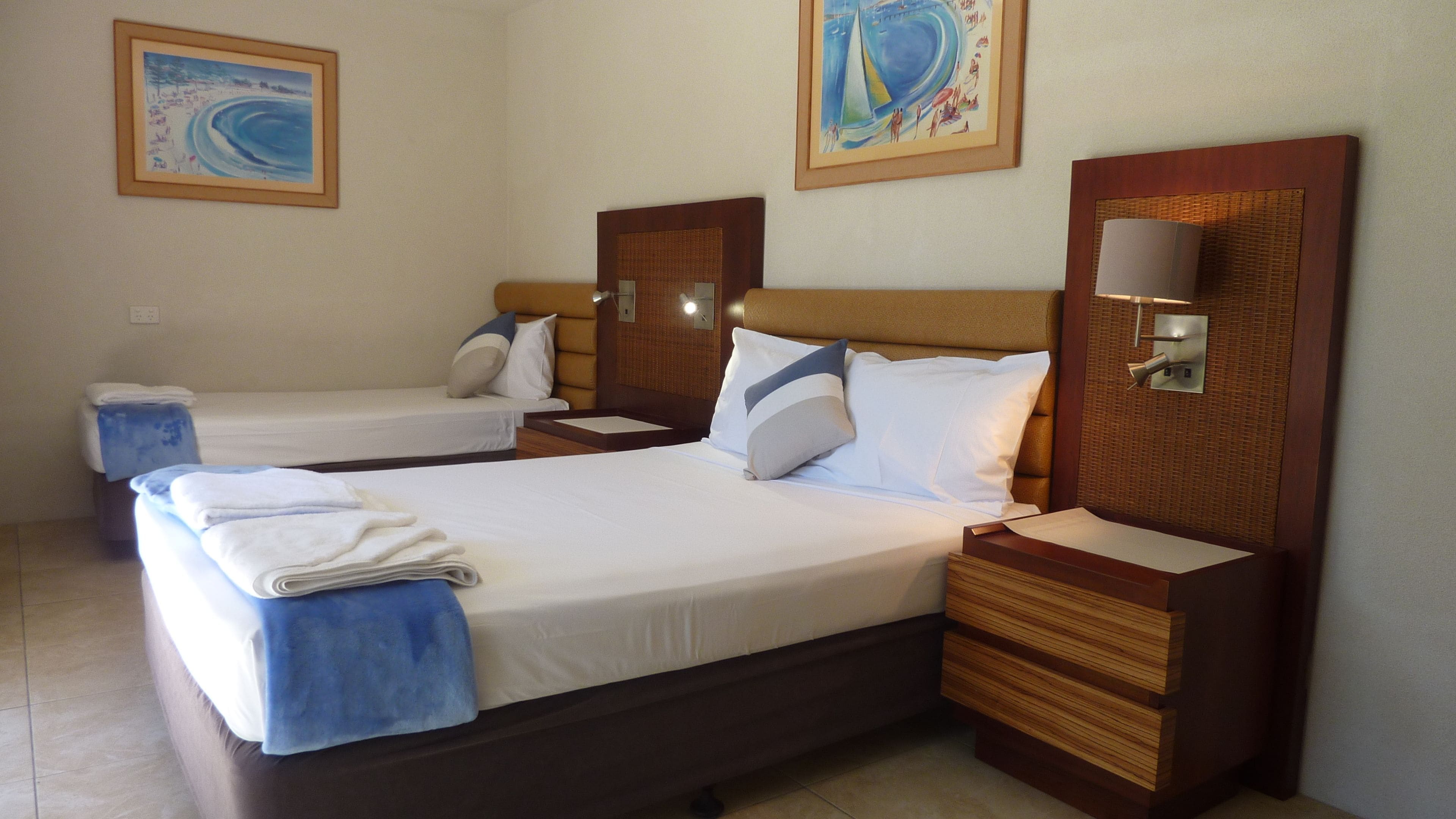 Habitación de hotel con dos camas, cada una con cabecero de madera, ropa de cama blanca y una toalla azul. Hay dos lámparas de pared y dos cuadros enmarcados sobre las camas.
