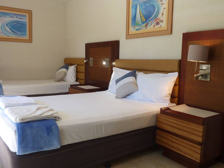 Habitación de hotel con dos camas, cada una con cabecero de madera, ropa de cama blanca y una toalla azul. Hay dos lámparas de pared y dos cuadros enmarcados sobre las camas.