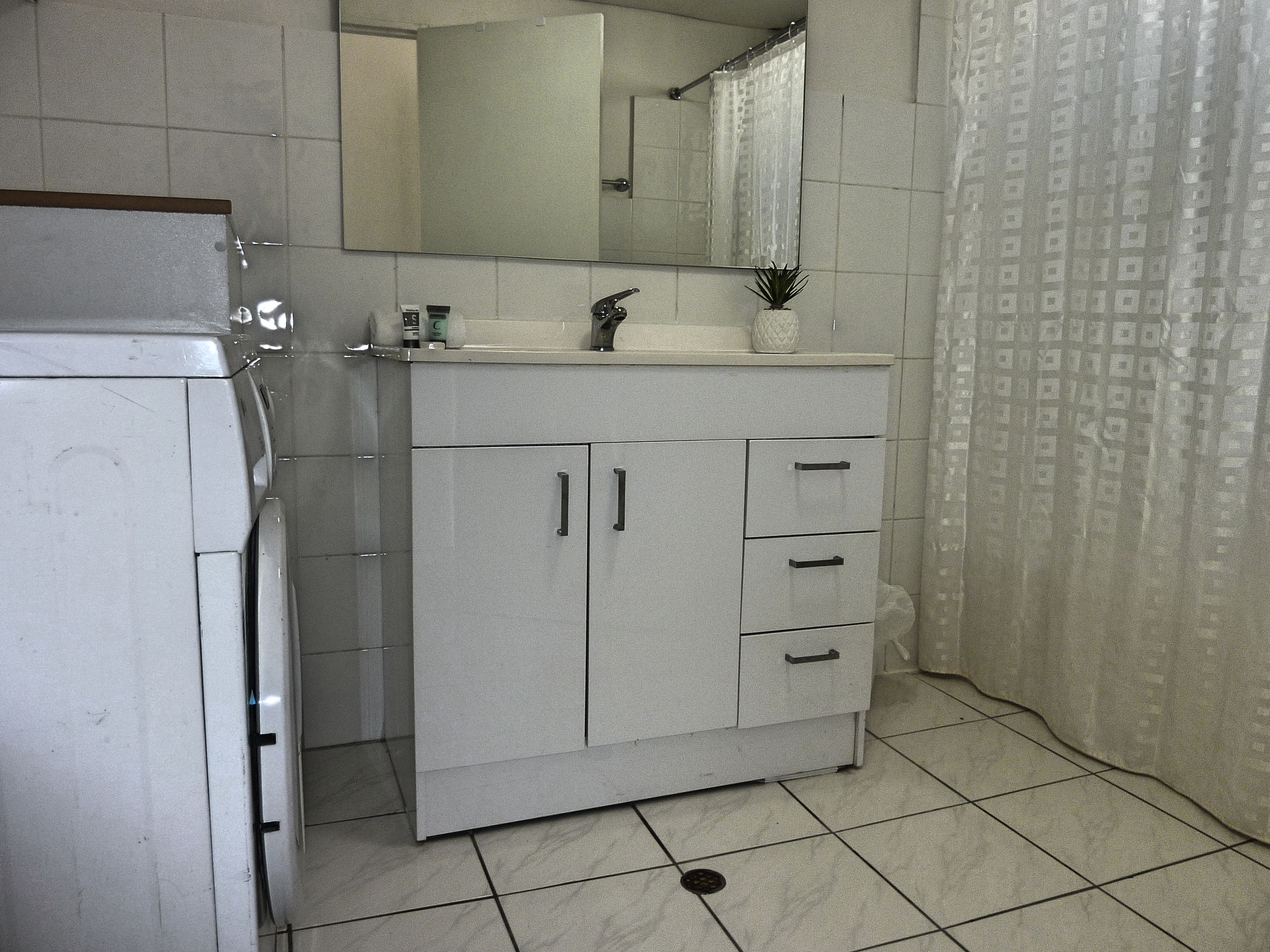 Un baño con lavamanos, espejo y mueble blanco, cortina de ducha y piso de baldosas.