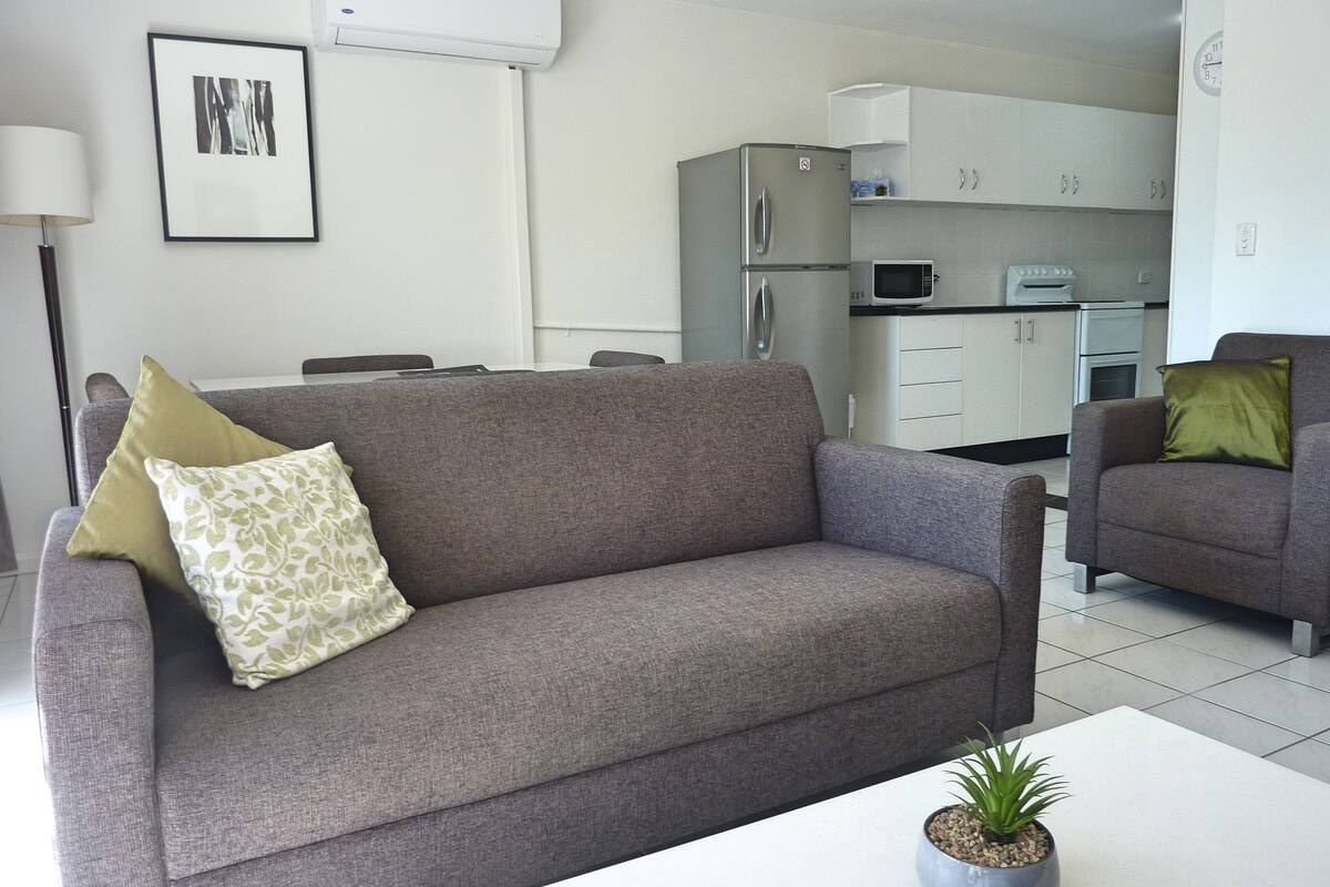 Una sala de estar con un sofá gris, una mesita con una planta y una cocina al fondo.