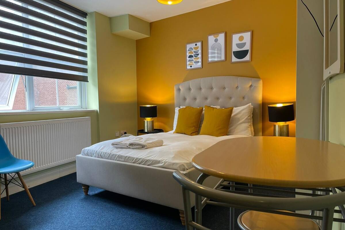 A hotel room with a bed, two bedside lamps, a round table, a chair, and artwork on the wall.