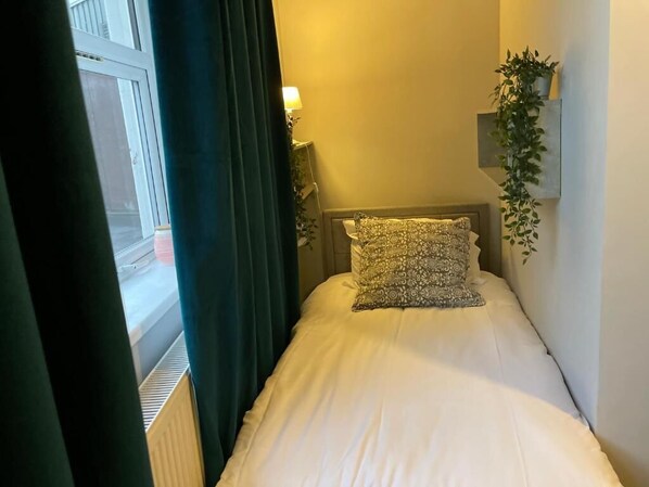 A hotel room with a bed, a nightstand with a lamp, a window with curtains, and a wall-mounted plant. - Brighter Properties (London)