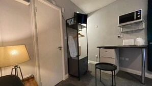 A compact hotel room with a kitchenette, a desk, a chair, a lamp, and a wardrobe. - Brighter Properties (London)
