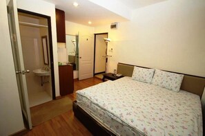A bedroom with a bed, a bedside table, a nightstand, a mirror, a door, and a bathroom with a sink and mirror.