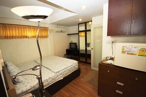 A compact hotel room with a bed, bedside lamp, a small desk with a television, a refrigerator, and a sliding door leading to a bathroom.