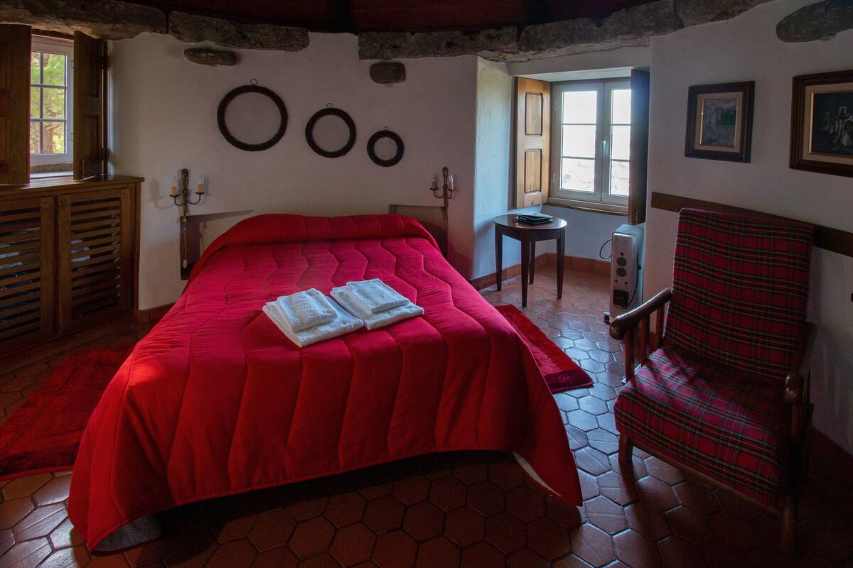 A room with a red bed, a wooden chair, a small table, and a window with a view.