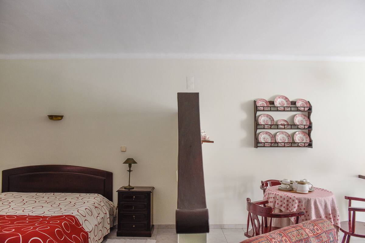 A bedroom with a bed, a nightstand, a lamp, a tall pillar, a dining table with chairs, and a wall-mounted dish rack.