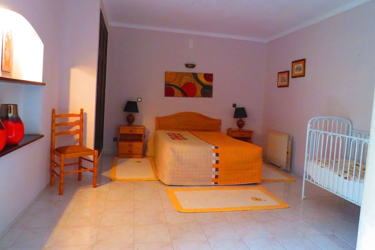 A bedroom with a bed, a chair, a nightstand, a lamp, a radiator, and a crib.