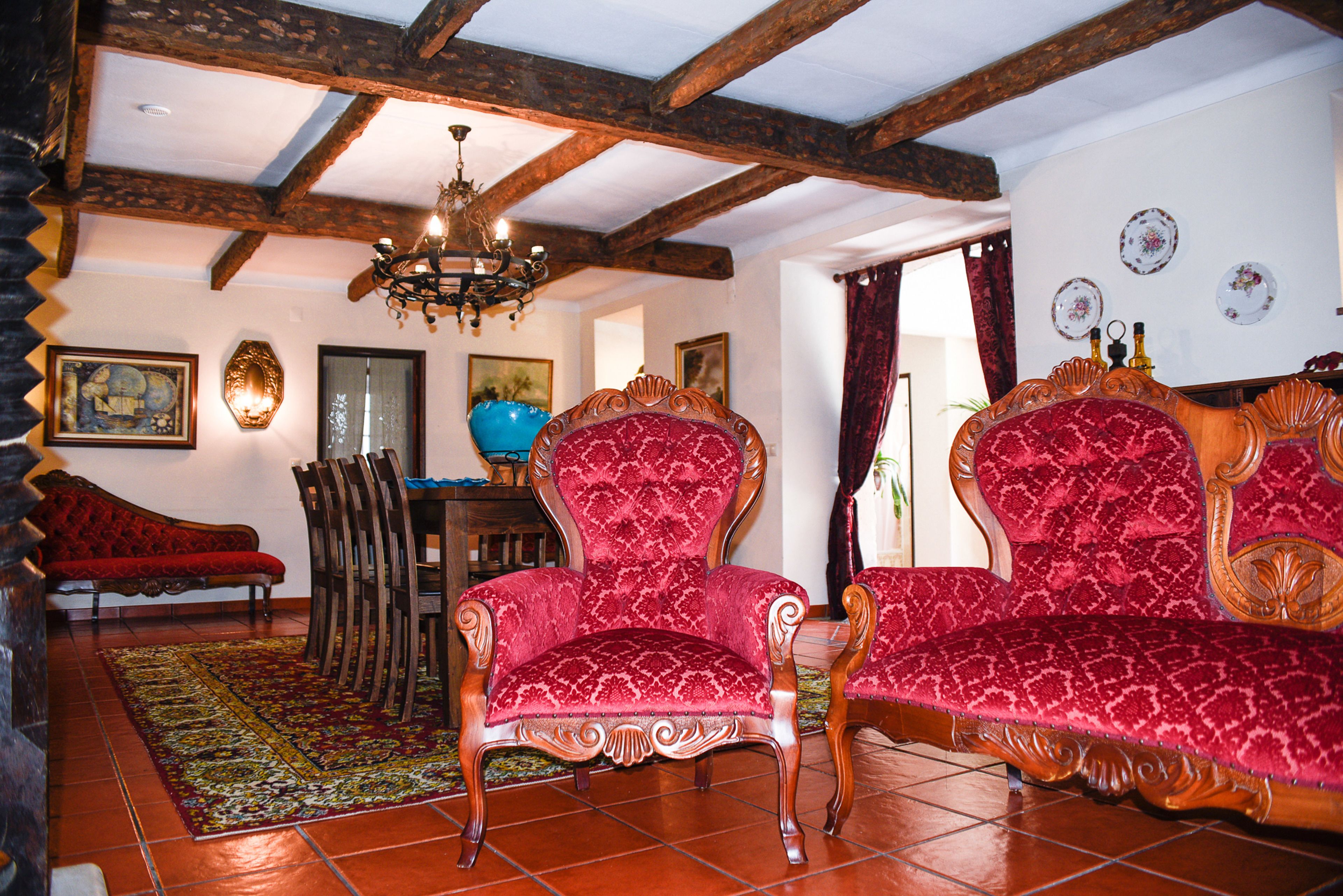 A traditional room with wooden beams, a chandelier, a red patterned sofa, a wooden chair, and a red sofa.