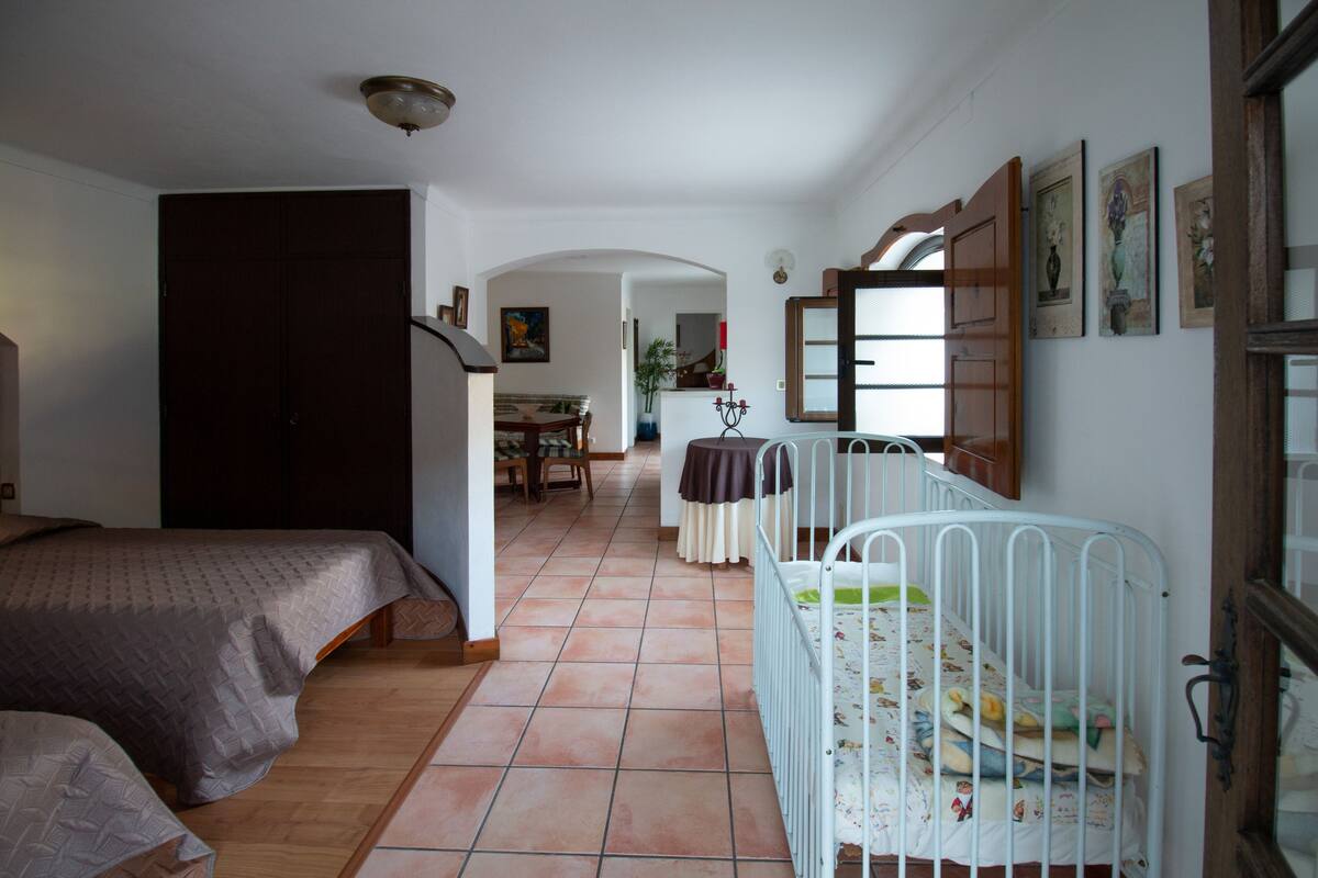 A bedroom with two beds, a crib, and a dining area in the background.