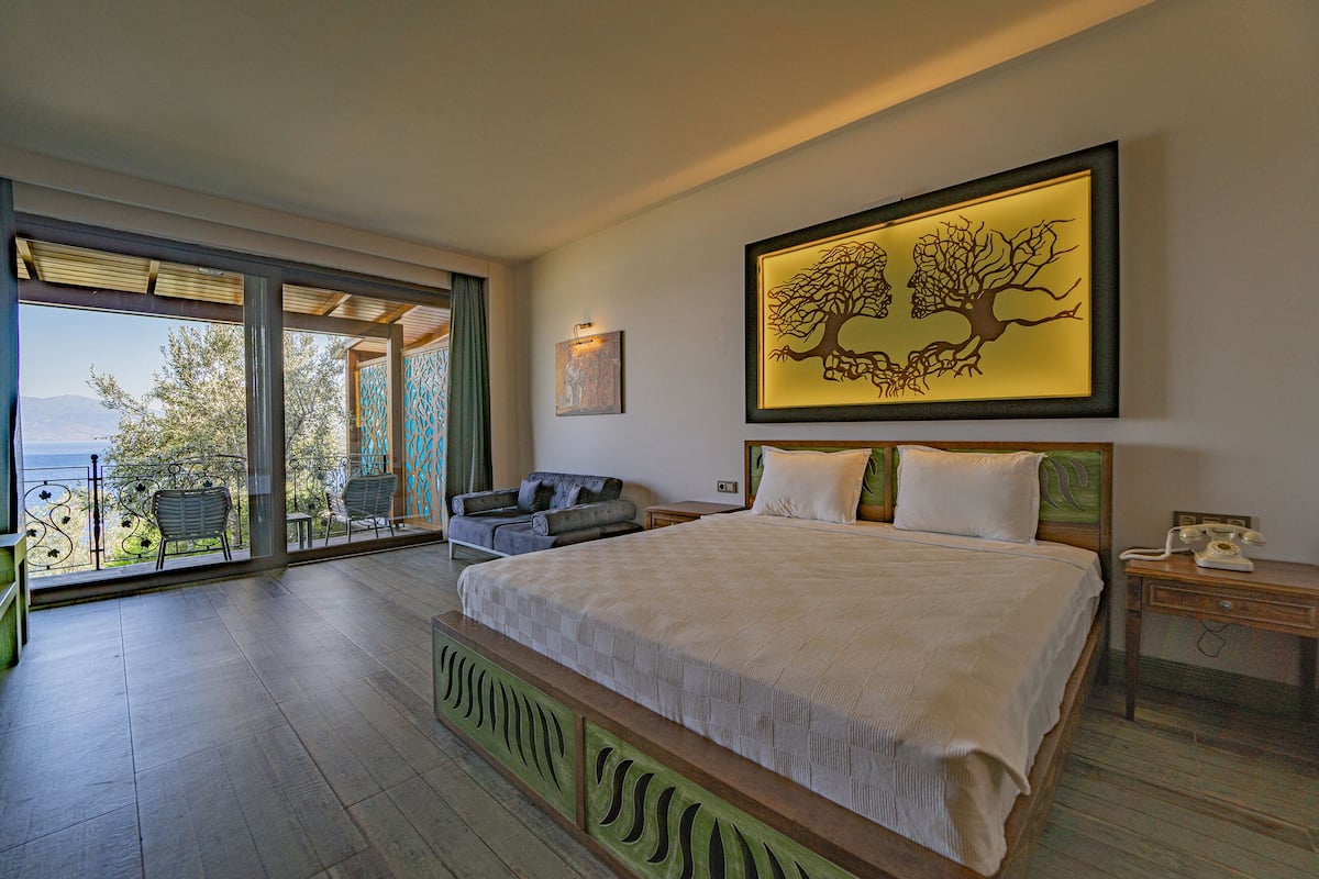 A bedroom with a large bed, a view through a glass door, a framed artwork, and a nightstand with a phone.