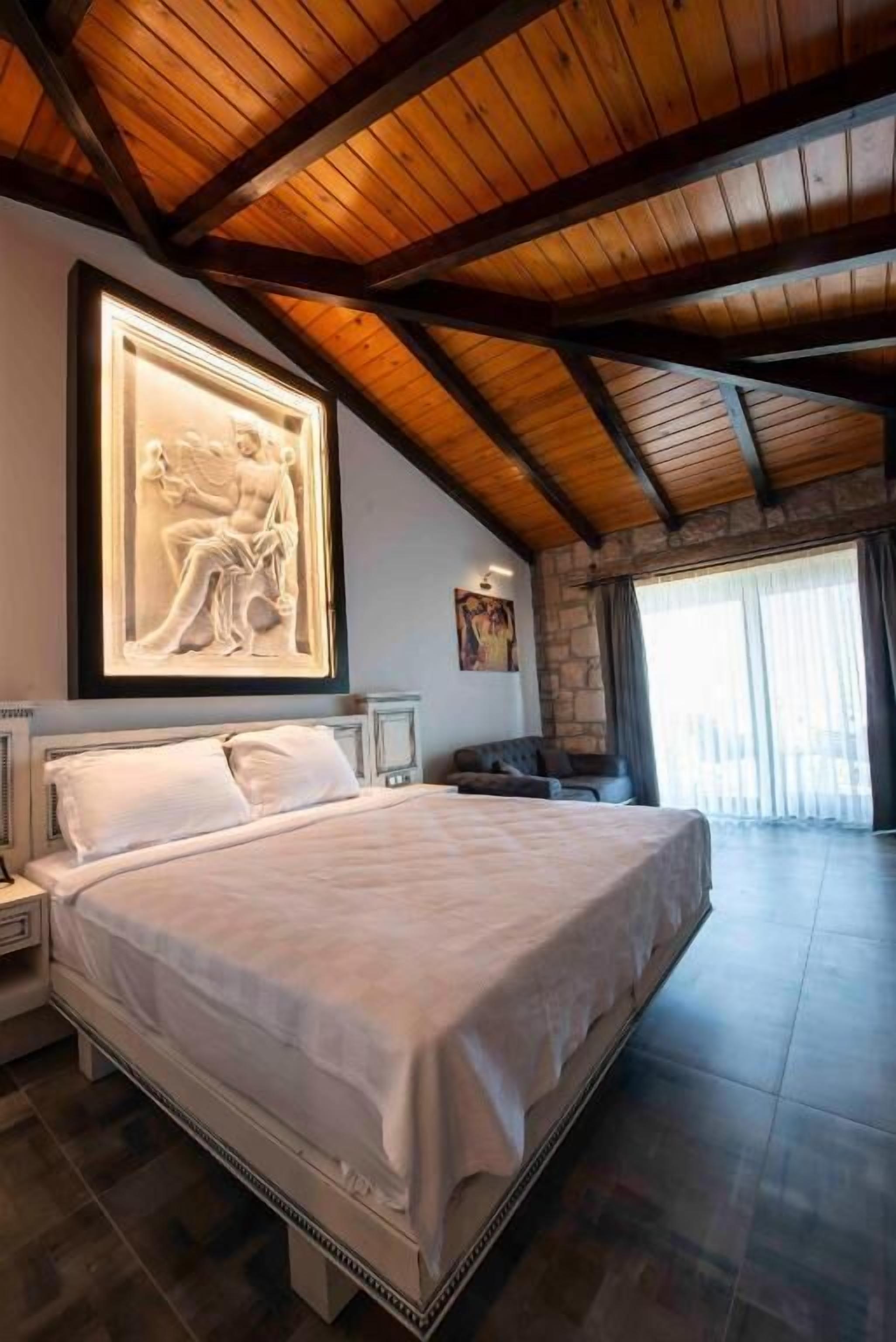A bedroom with a large bed, a wooden ceiling, a stone wall, and a large window.