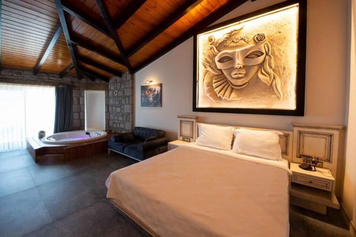 A bedroom with a large bed, a sitting area with a chair, a bathtub, and a wall-mounted artwork.