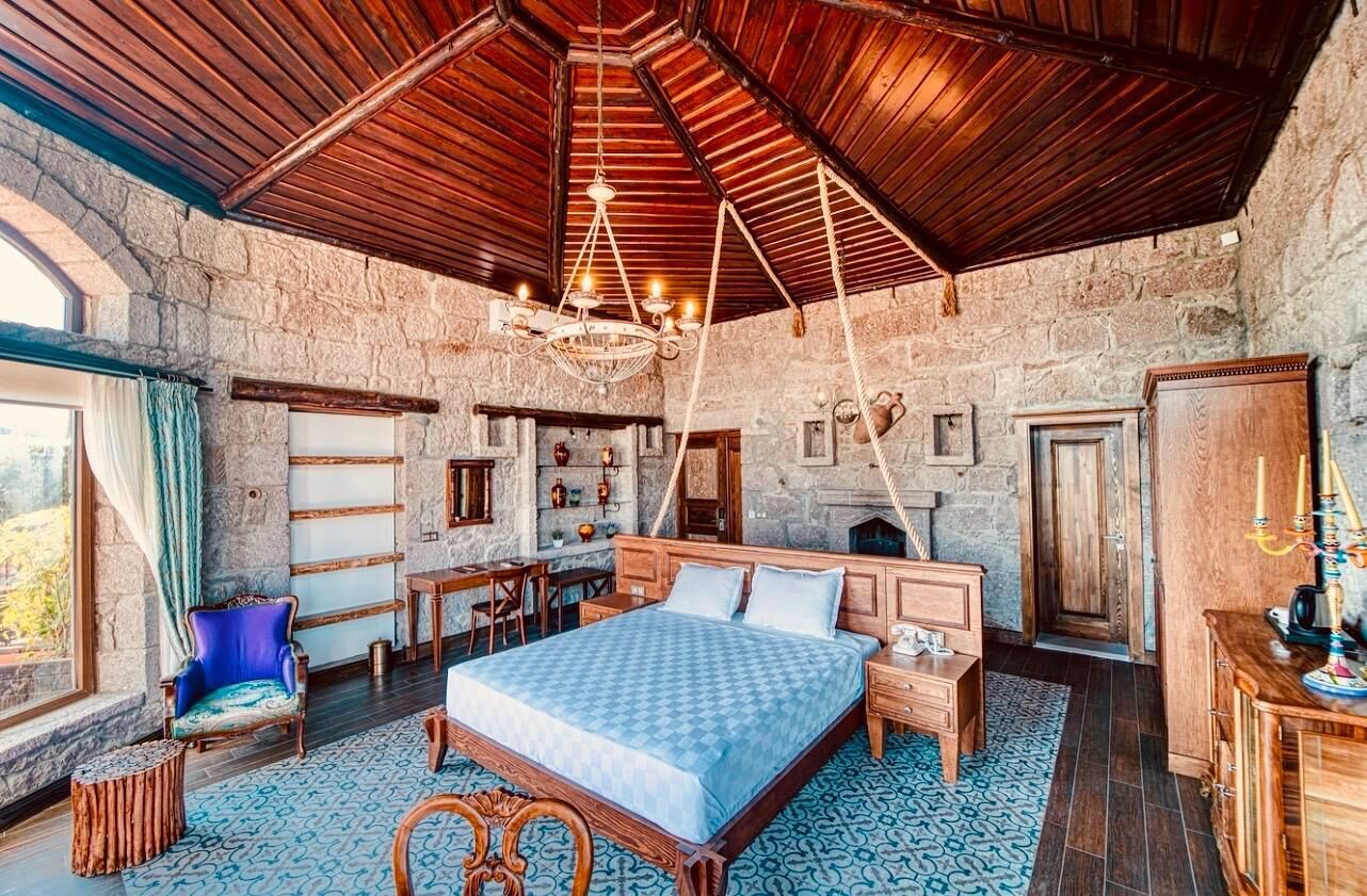 A bedroom with a large bed, wooden ceiling, stone walls, a chandelier, and a view through a window.