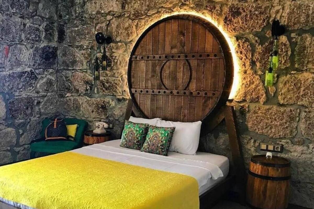 A bed with a yellow bedspread, a stone wall, a wooden barrel, and a green chair.