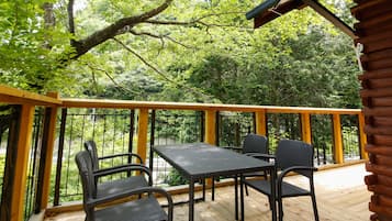 Una terraza de madera con una mesa y sillas, rodeada por una barandilla metálica, con vistas a un bosque frondoso.