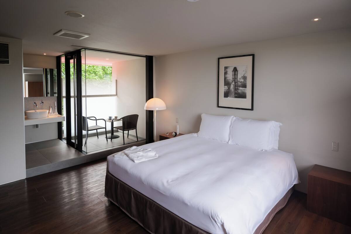 A modern hotel room with a large bed, a bathroom with a sink and mirror, a chair, and a framed picture on the wall.