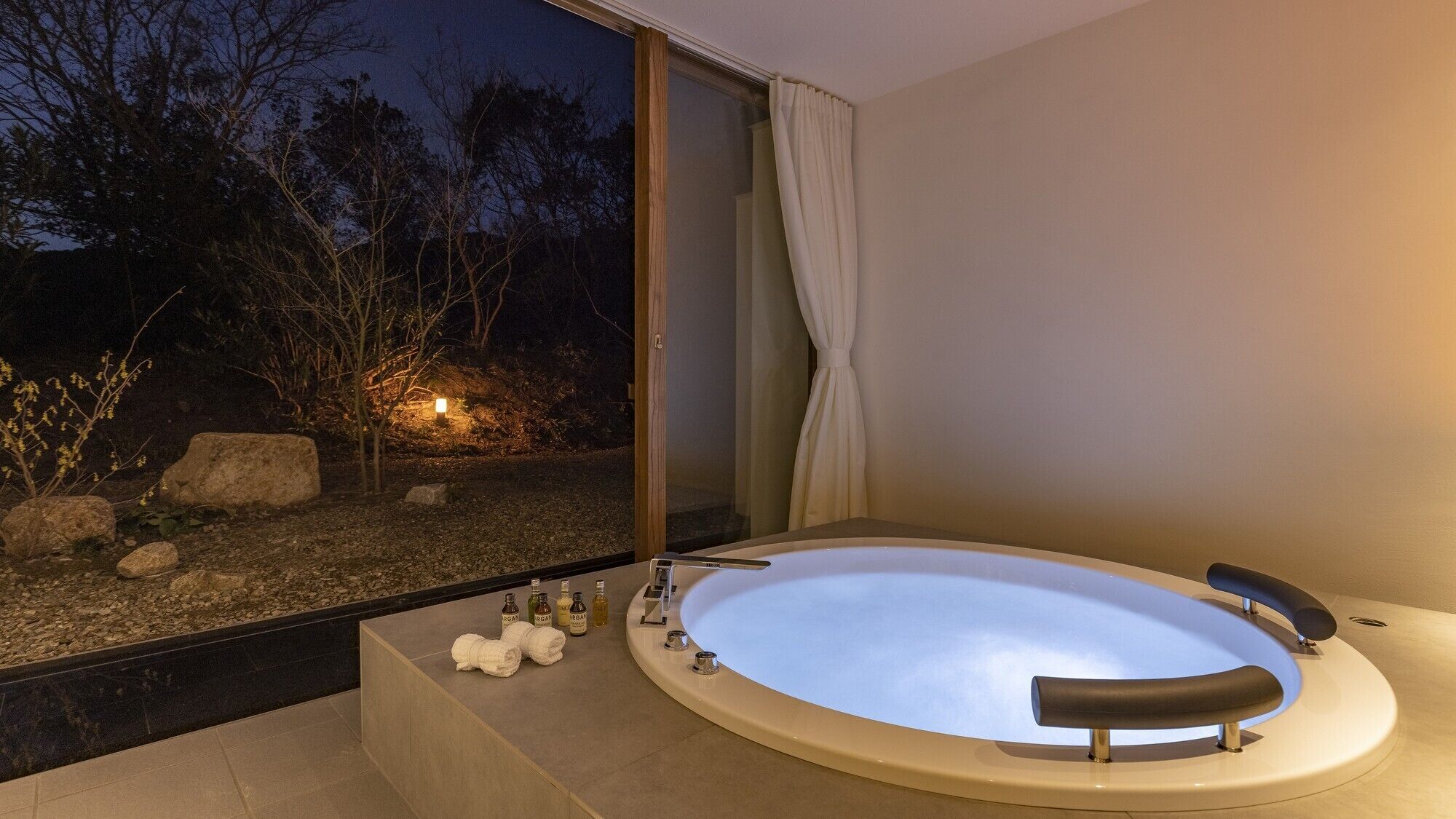 Un baño moderno con una bañera grande, cabina de ducha de vidrio y vistas a un jardín por la noche.