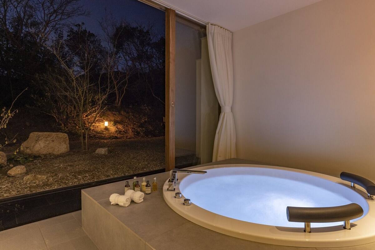A modern bathroom with a large bathtub, a glass shower enclosure, and a view of a garden at night.