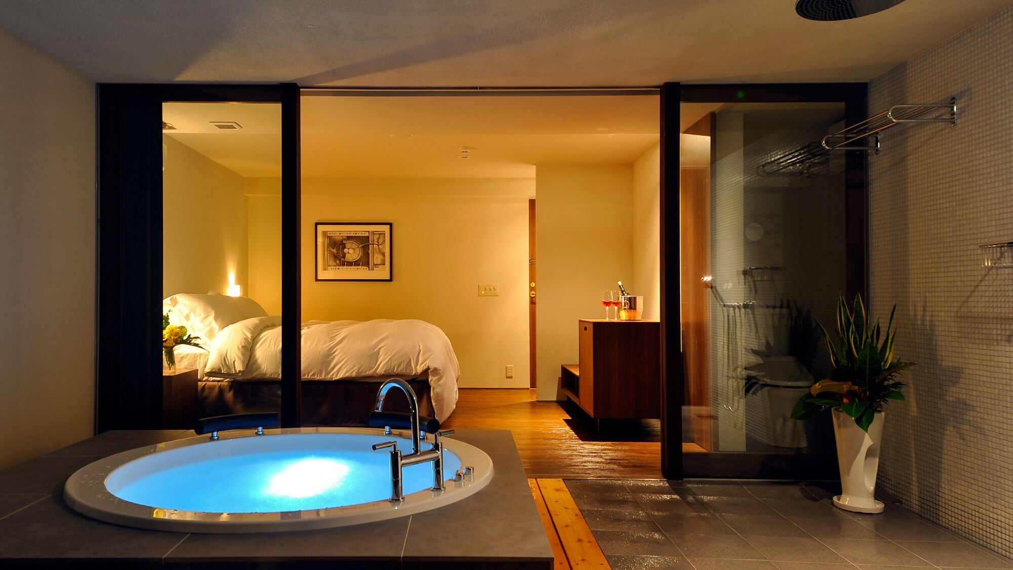 Un baño moderno con bañera, zona de ducha y un dormitorio visible con cama y mesita de noche.