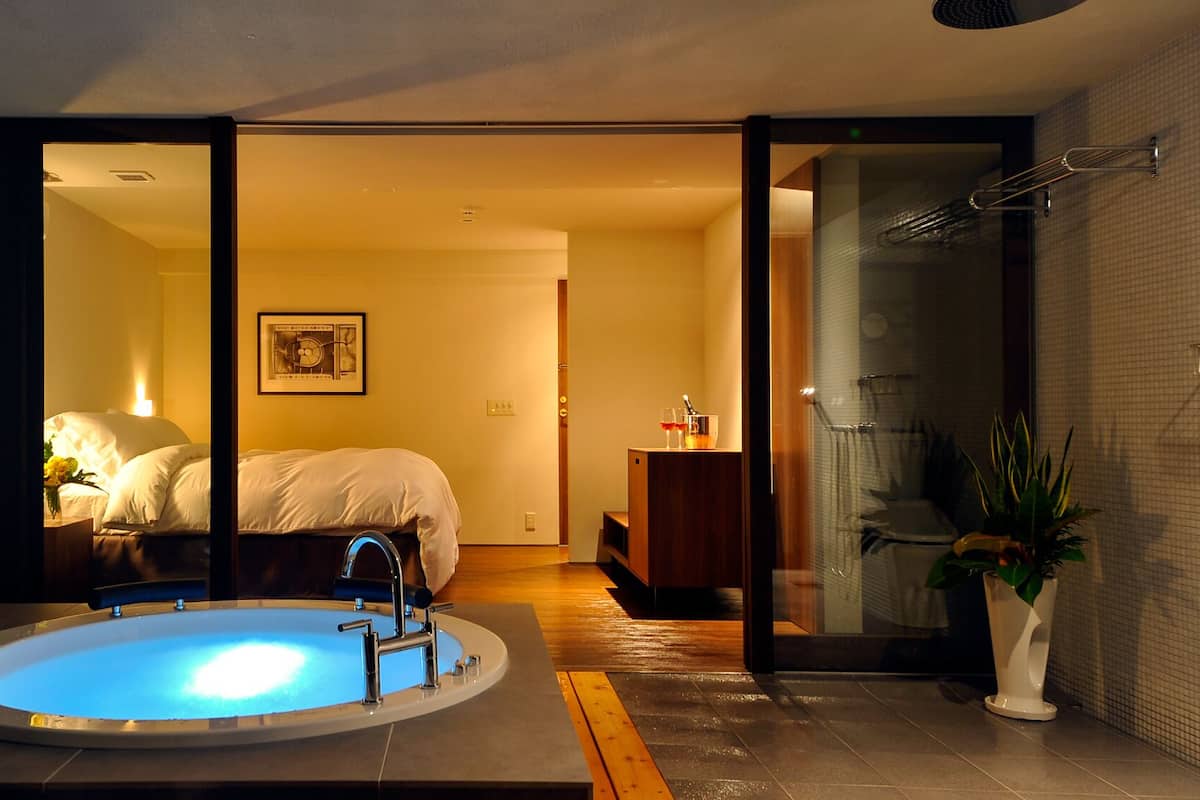 A modern bathroom with a bathtub, a shower area, and a visible bedroom with a bed and a nightstand.