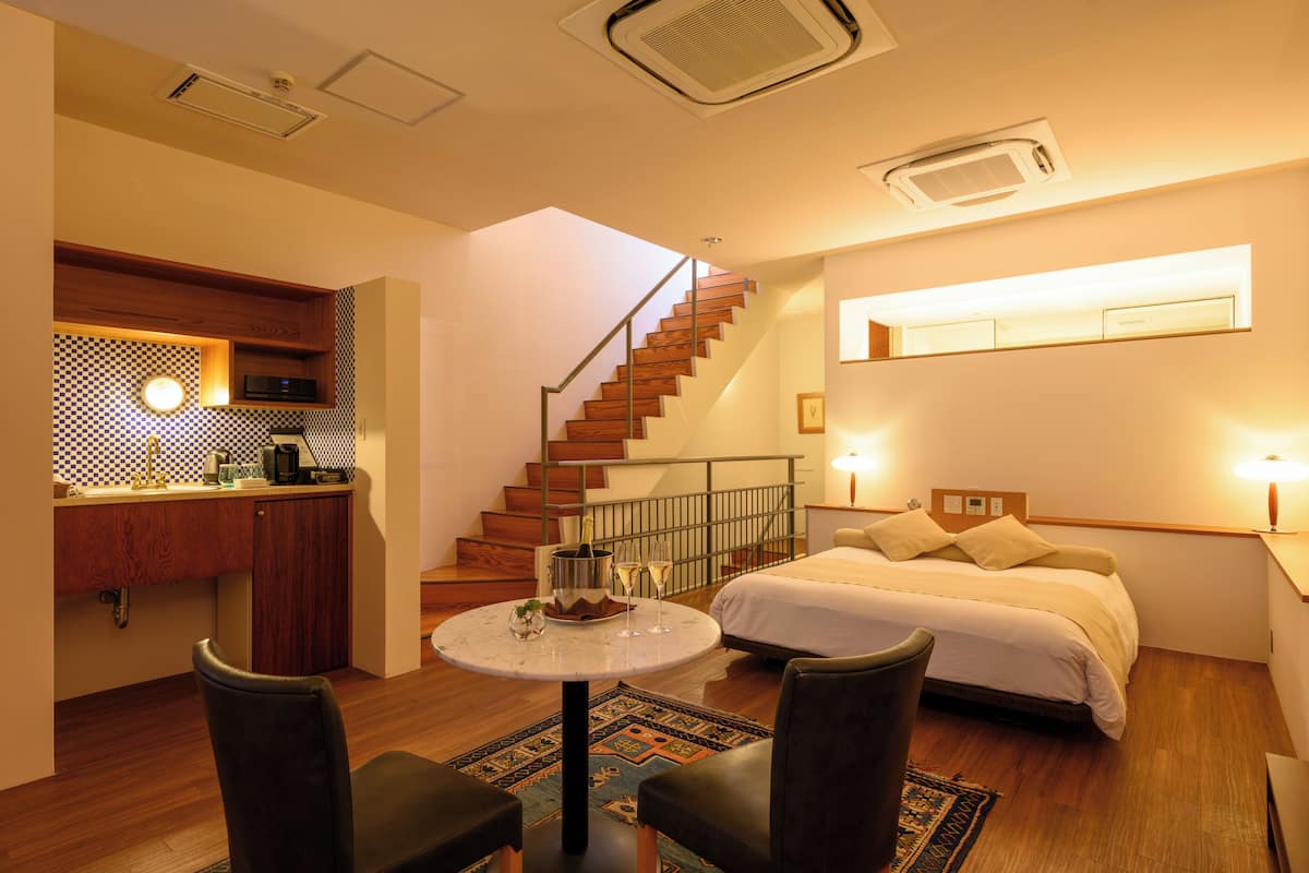 A compact hotel room with a bed, a small dining table, two chairs, a kitchen area with a sink, and a staircase.