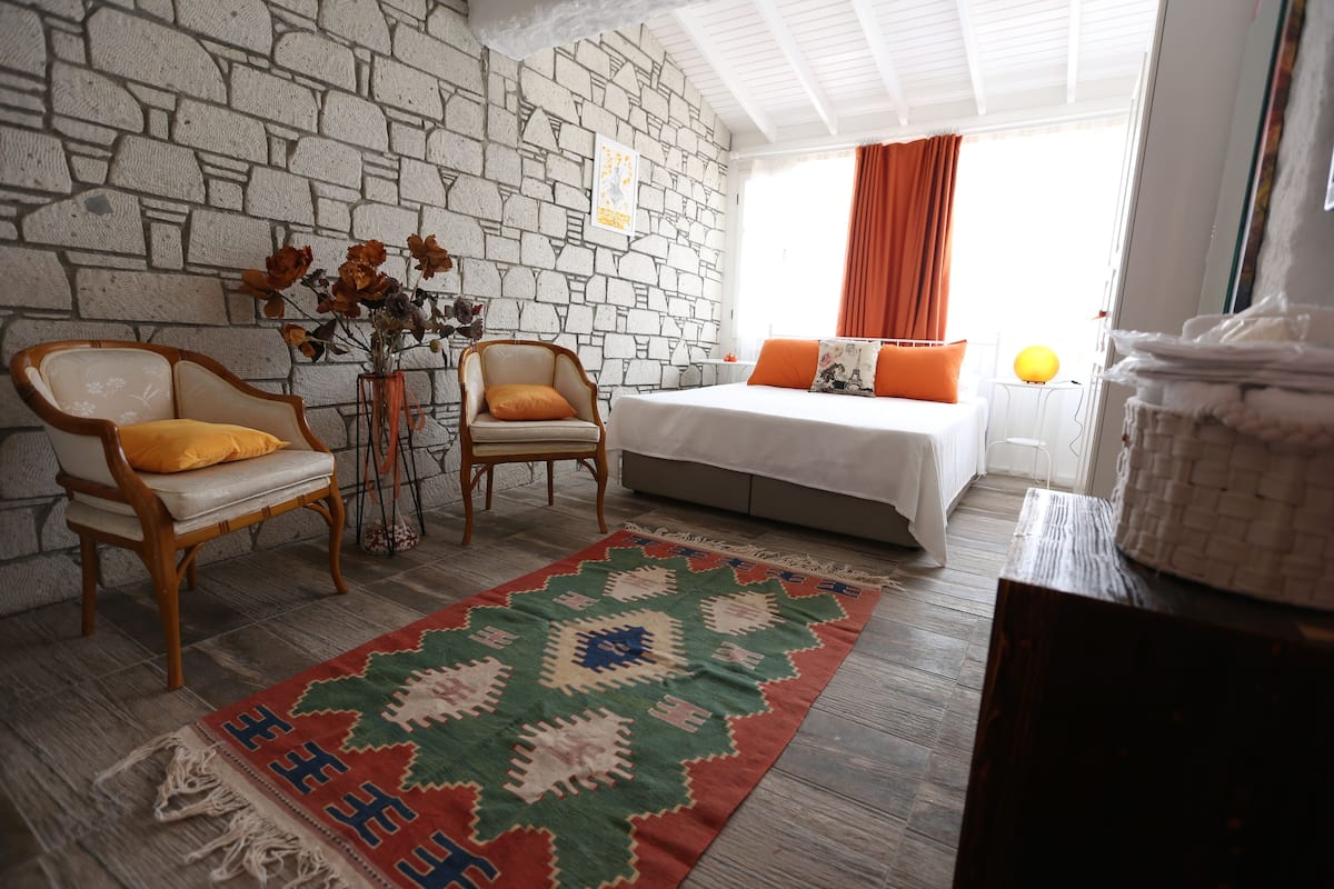 A room with a stone wall, a bed with orange pillows, a wooden chair with a yellow cushion, and a patterned rug.
