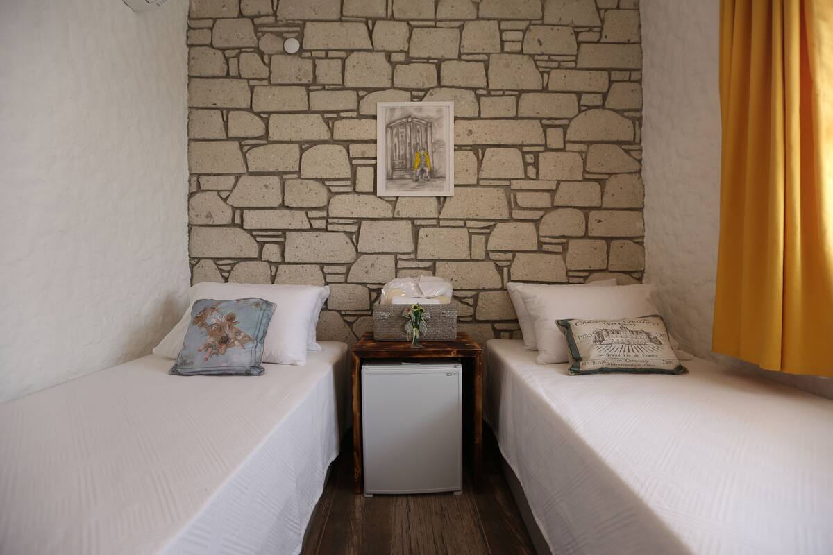 A room with two single beds, a small wooden nightstand with a refrigerator, and a stone wall.