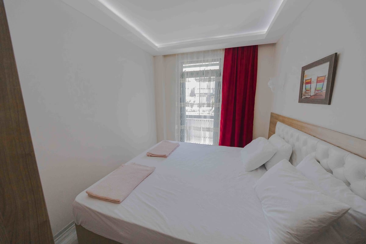 A hotel room with a large bed, a wooden headboard, a window with red curtains, and a framed picture on the wall.