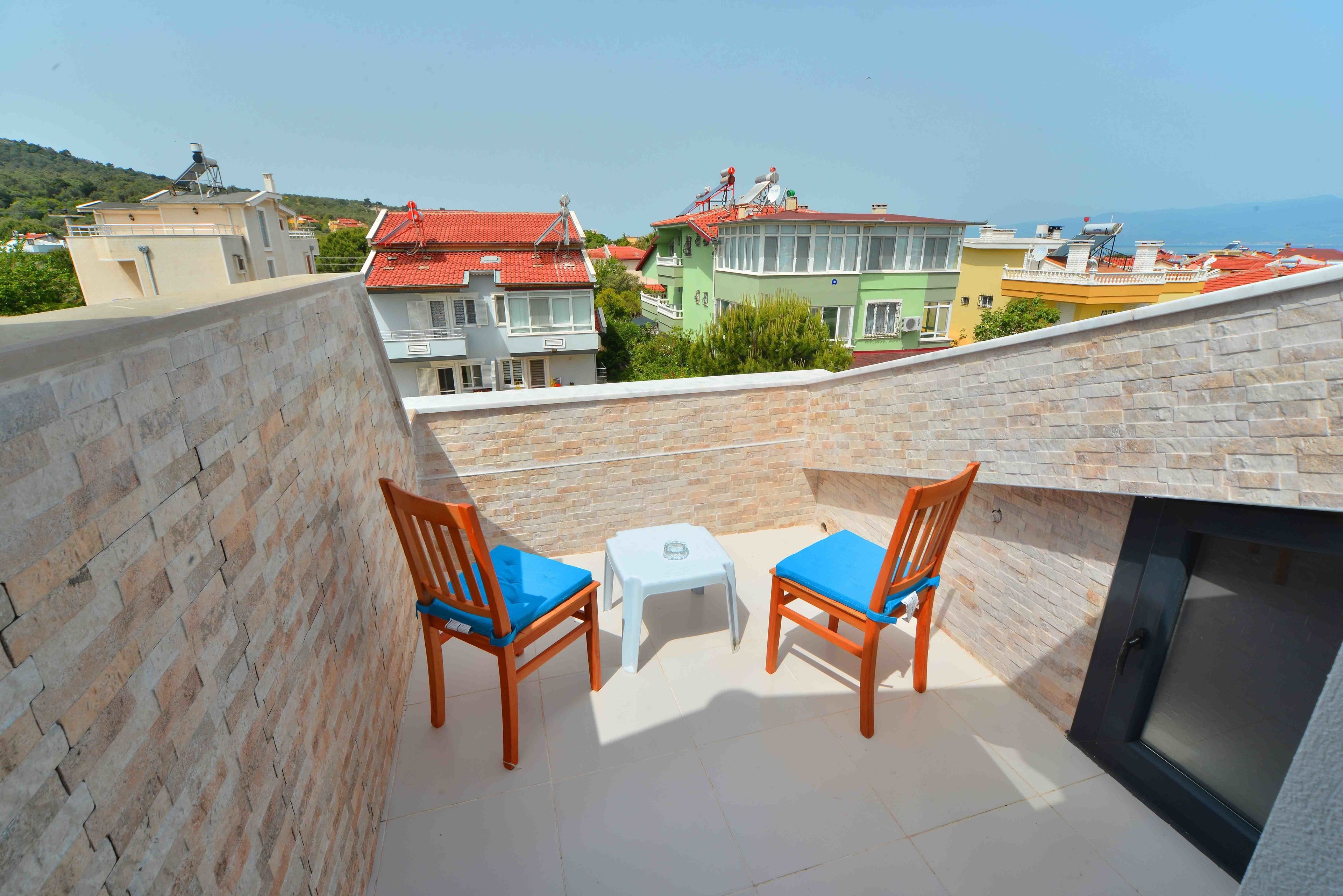 A balcony with two wooden chairs and a small table, overlooking colorful buildings and a distant view.