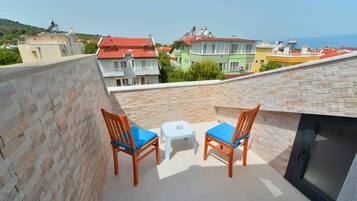 A balcony with two wooden chairs and a small table, overlooking colorful buildings and a distant view.
