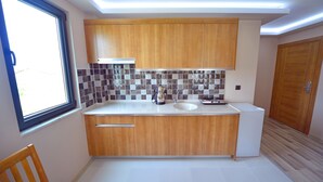 A compact kitchen with wooden cabinets, a white countertop, a built-in sink, and a window providing natural light. - Asil Apart Otel (Burhaniye)