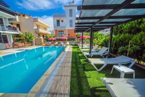 A swimming pool with lounge chairs and umbrellas, surrounded by a well-maintained lawn and buildings in the background. - Asil Apart Otel (Burhaniye)