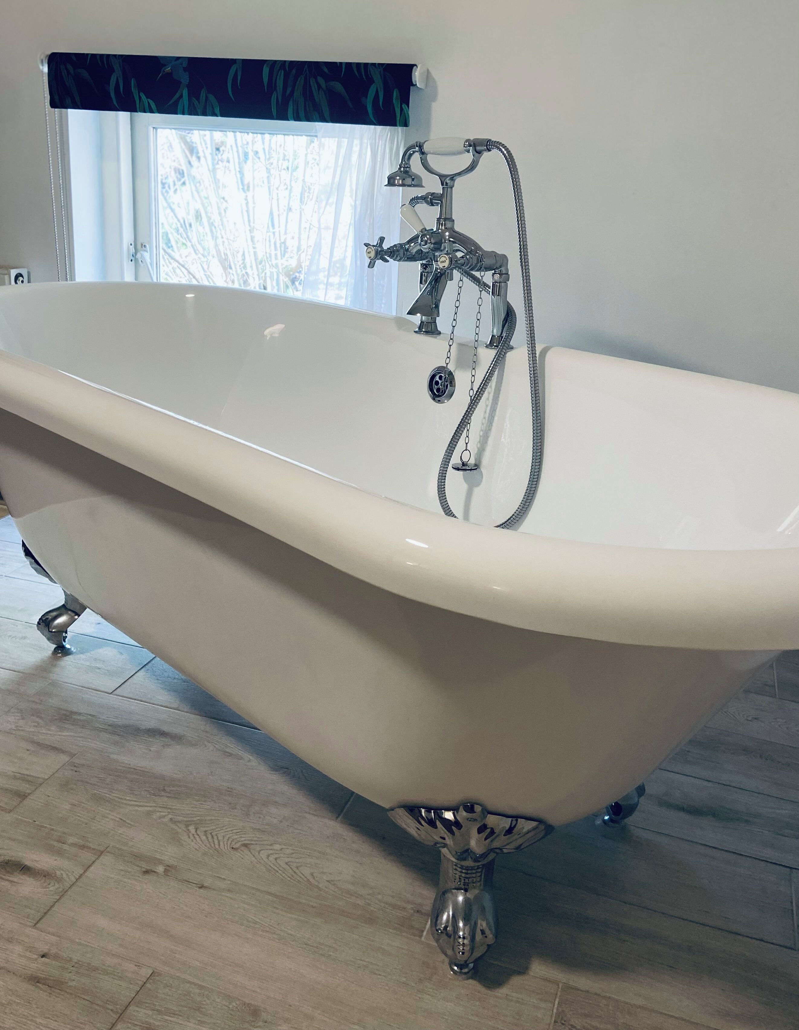 A clawfoot bathtub with a shower attachment in a room with wooden flooring and a window with curtains.