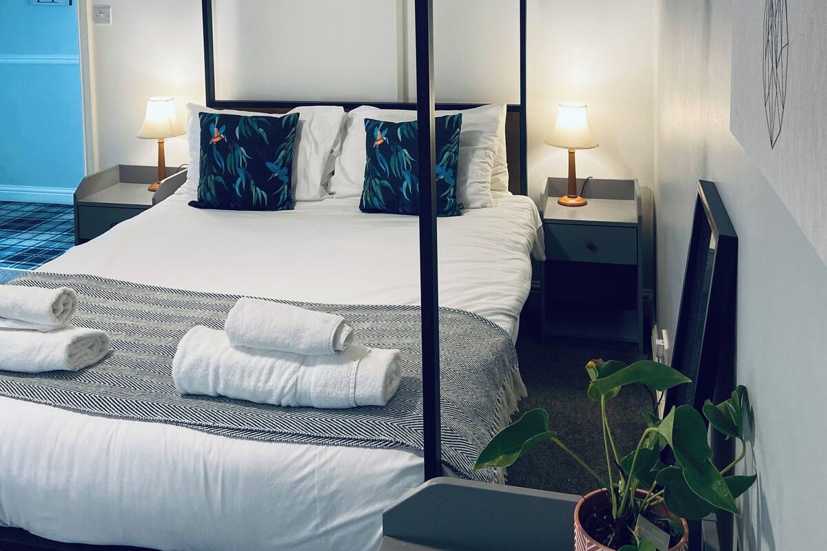 A four-poster bed with white linens and blue patterned pillows, two bedside lamps, a potted plant, and a framed picture on the wall.