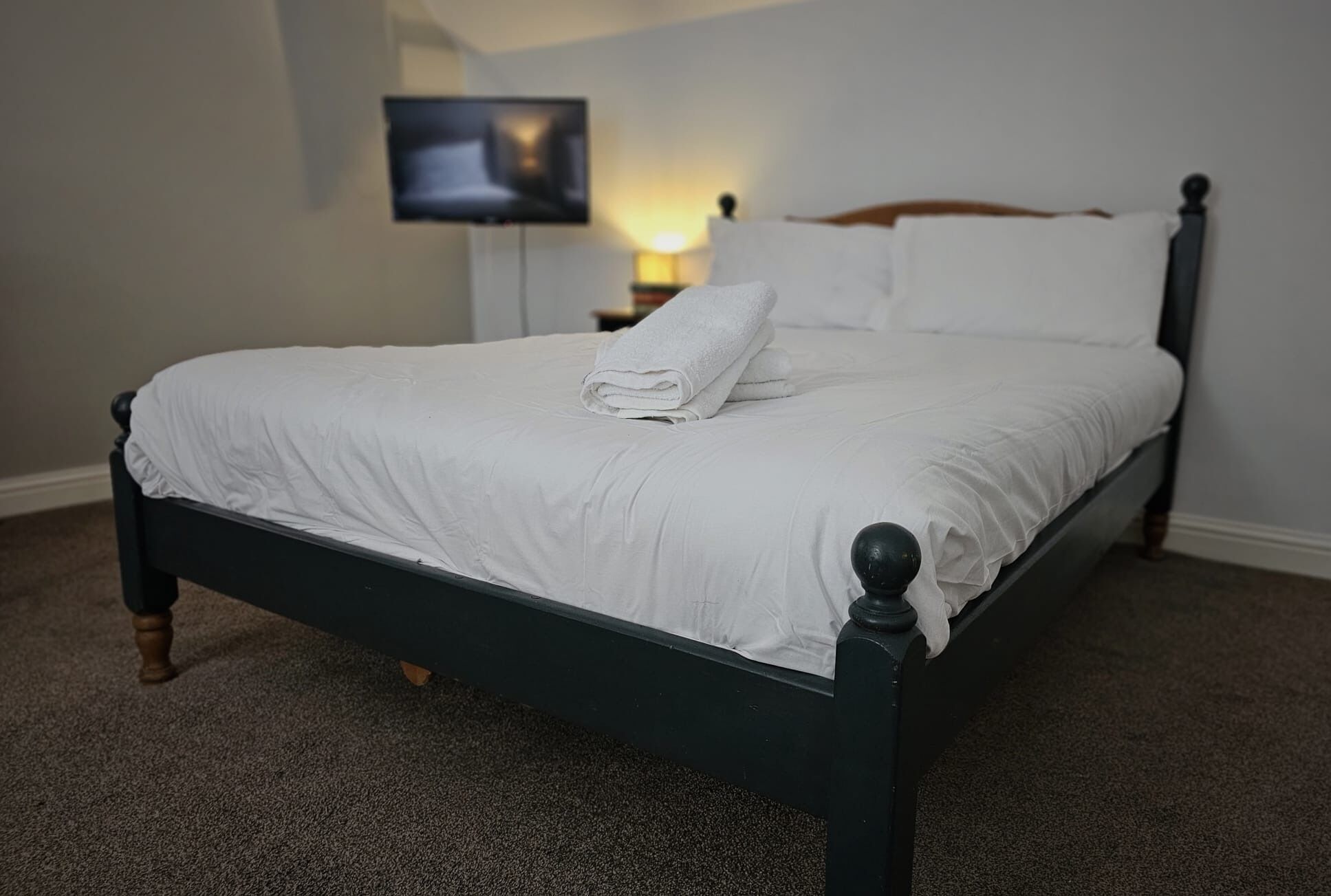 A neatly made bed with white linens and a folded towel, a dark wooden headboard, and a television mounted on the wall in the background.