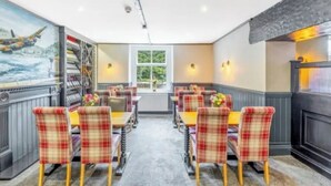A restaurant with a central dining area featuring plaid chairs and tables, a mural on the left wall, and a window with a view of greenery. - Ladybower Inn (Hope Valley)