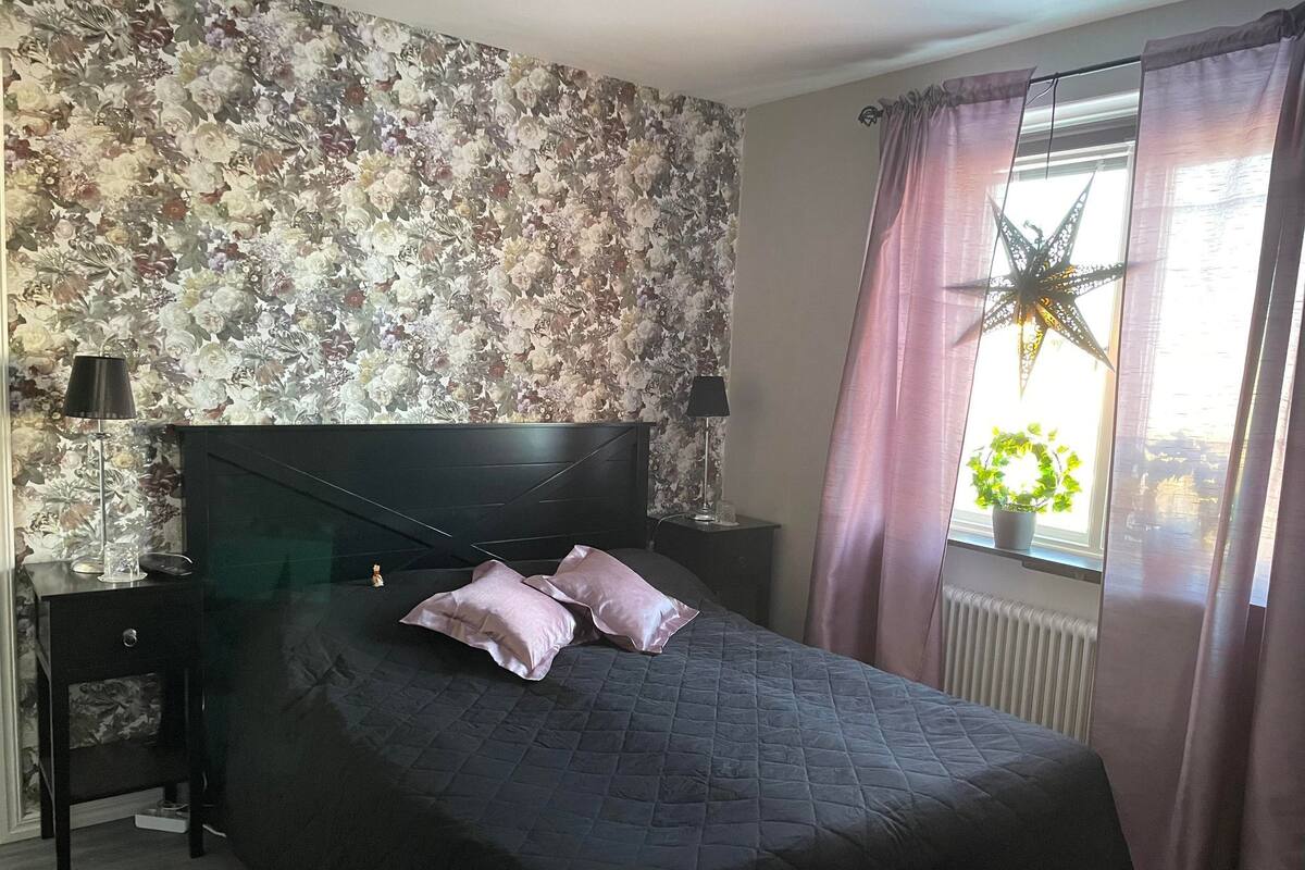 Een slaapkamer met bloemenbehang, een zwart hoofdeinde, een bed met quilt, twee bedlampen, een nachtkastje, een raam met gordijnen en een ster decoratie.